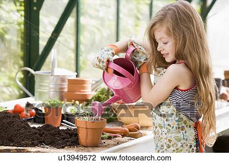 Stock Image - Young girl in greenhouse watering potted plant smiling. Fotosearch