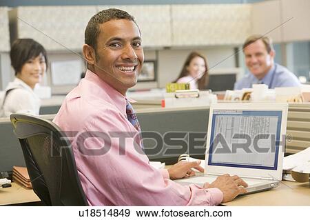 Stock Photo - Businessman in cubicle using laptop and smiling. Fotosearch