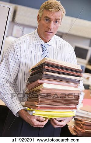 Stock Image - Businessman standing in cubicle with stacks of files. Fotosearch