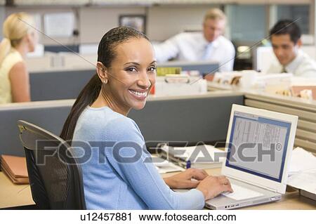 Stock Image - Businesswoman in cubicle using laptop smiling. Fotosearch