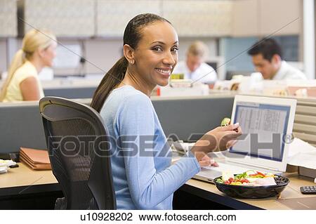 Stock Image - Businesswoman in cubicle with laptop eating salad. Fotosearch