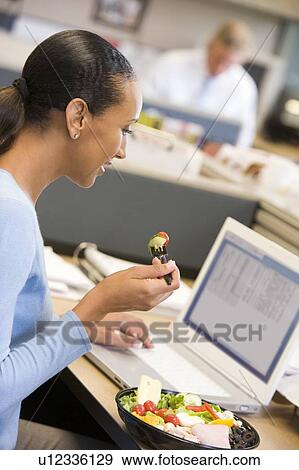 Stock Photo - Businesswoman in cubicle with laptop eating salad. Fotosearch