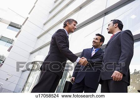Picture - Three businessmen standing outdoors by building shaking hands and smiling (high key/selective focus). Fotosearch
