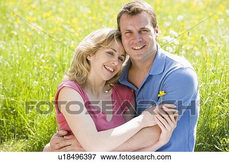 Stock Photo - Couple sitting outdoors holding flower smiling. Fotosearch