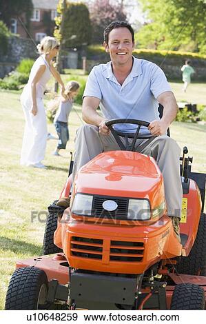 Stock Photo - Man outdoors driving lawnmower smiling with family in background. Fotosearch