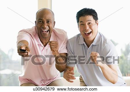 Stock Photo - Two men in living room with remote control cheering and smiling. Fotosearch