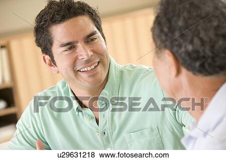 Stock Photo - Two men sitting in living room talking and smiling. Fotosearch