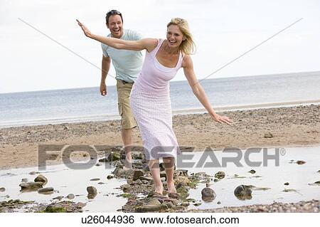 Stock Photograph - Couple at the beach walking on stones and smiling. Fotosearch