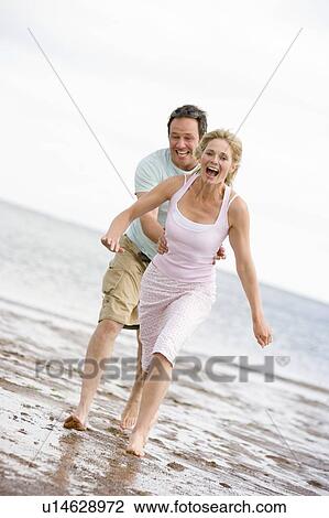 Stock Image - Couple running at the beach smiling. Fotosearch