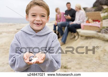 Stock Image - Family at beach with picnic smiling focus on boy with seashells. Fotosearch