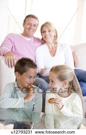 Stock Image - Family sitting in living room eating cookies and smiling. Fotosearch