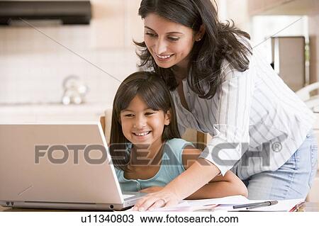 Stock Image - Woman and young girl in kitchen with laptop and paperwork smiling. Fotosearch