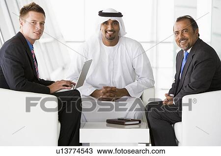 Stock Image - Three businessmen indoors with a laptop smiling (high key/selective focus). Fotosearch