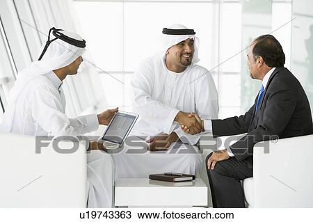 Stock Image - Three businessmen indoors with a laptop shaking hands and smiling (high key/selective focus). Fotosearch