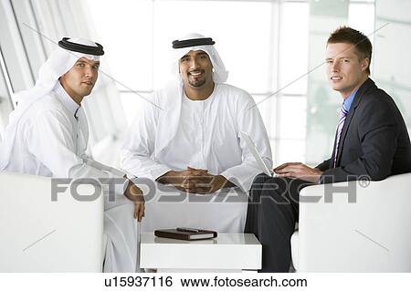 Stock Photograph - Three businessmen indoors with a laptop smiling (high key/selective focus). Fotosearch