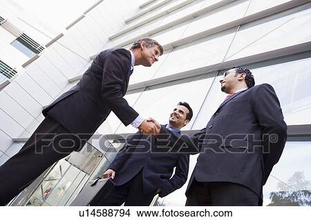 Three businessmen standing outdoors by building shaking hands and smiling (high key/selective focus) View Large Photo Image Picture - Three businessmen standing outdoors by building shaking hands and smiling (high key/selective focus). Fotosearch