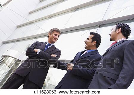 Stock Image - Three businessmen standing outdoors by building talking and smiling (high key/selective focus). Fotosearch