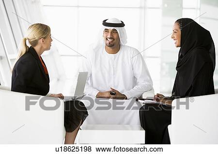 Stock Photo - Three businesspeople indoors with a laptop and cellular phone talking and smiling (high key/selective focus). Fotosearch