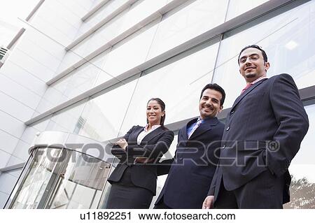 Stock Image - Three businesspeople standing outdoors by building smiling (high key/selective focus). Fotosearch