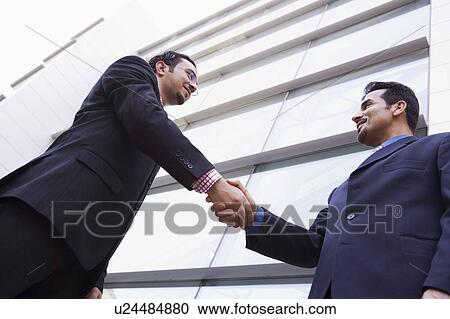 Stock Image - Two businessmen outdoors by building shaking hands and smiling (high key/selective focus). Fotosearch
