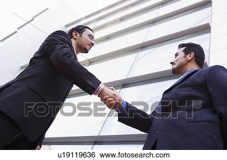 Stock Photograph - Two businessmen outdoors by building shaking hands (high key/selective focus). Fotosearch