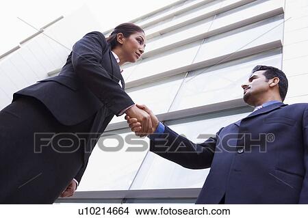 Picture - Two businesspeople outdoors by building shaking hands (high key/selective focus). Fotosearch