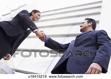 Stock Photo - Two businesspeople outdoors by building shaking hands and smiling (high key/selective focus). Fotosearch