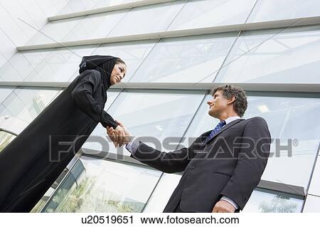 Stock Image - Two businesspeople standing outdoors by building shaking hands and smiling (selective focus). Fotosearch