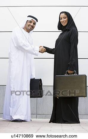 Stock Photo - Two businesspeople standing outdoors with briefcases shaking hands and smiling. Fotosearch