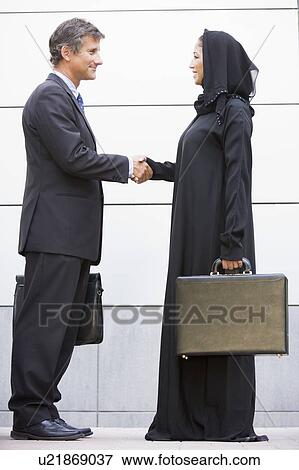 Stock Photo - Two businesspeople standing outdoors with briefcases shaking hands and smiling. Fotosearch