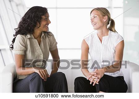 Stock Image - Two businesswomen sitting indoors talking and smiling (high key/selective focus). Fotosearch