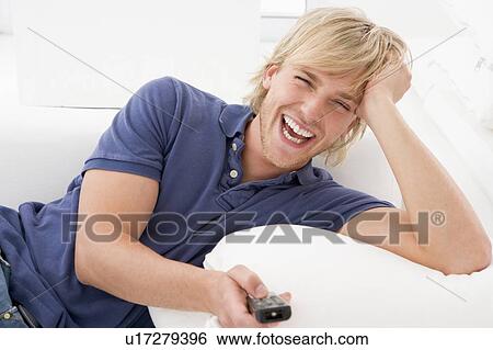 Stock Photograph - Man in living room holding remote control laughing. Fotosearch