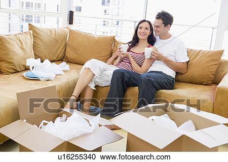 Stock Image - Couple relaxing with coffee by boxes in new home smiling. Fotosearch