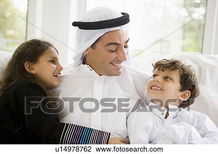 Stock Image - Father and two young children sitting in living room smiling (high key/selective focus). Fotosearch