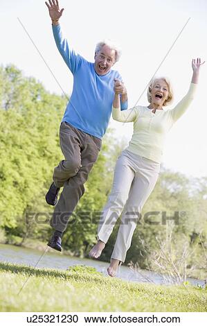 Stock Image - Couple jumping outdoors at park by lake smiling. Fotosearch