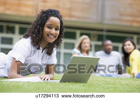 Stock Image - Student outdoors on lawn using laptop with other students in background (selective focus). Fotosearch