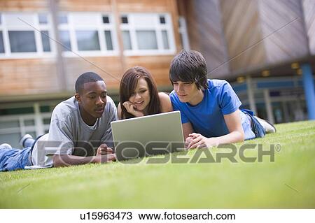Stock Image - Three students lying outdoors on lawn with laptop. Fotosearch