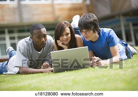 Stock Image - Three students lying outdoors on lawn with laptop. Fotosearch