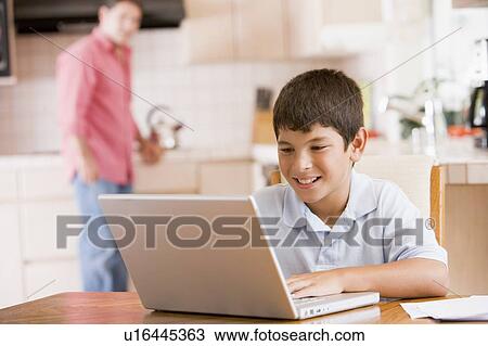 Stock Image - Young boy in kitchen with laptop and paperwork smiling with man in background. Fotosearch
