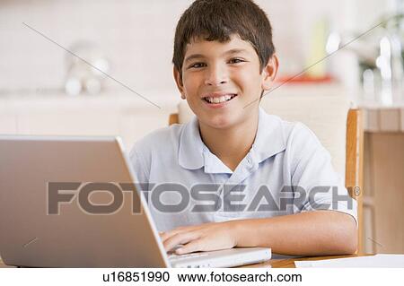 Stock Image - Young boy in kitchen with laptop and paperwork smiling. Fotosearch