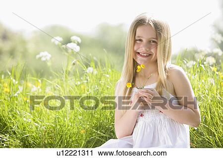 Stock Image - Young girl sitting outdoors holding flower smiling. Fotosearch