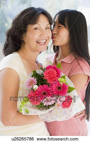Stock Photograph - Granddaughter kissing grandmother on cheek holding flowers and smiling. Fotosearch