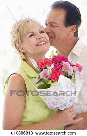 Stock Image - Husband and wife holding flowers kissing and smiling. Fotosearch