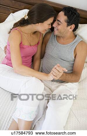 Stock Image - Couple relaxing on bed holding hands in bedroom smiling (selective focus). Fotosearch