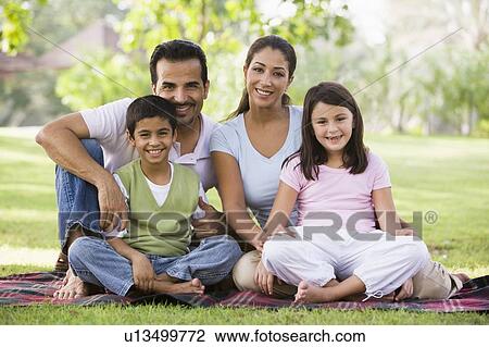 Stock Image - Family sitting outdoors in park smiling (selective focus). Fotosearch