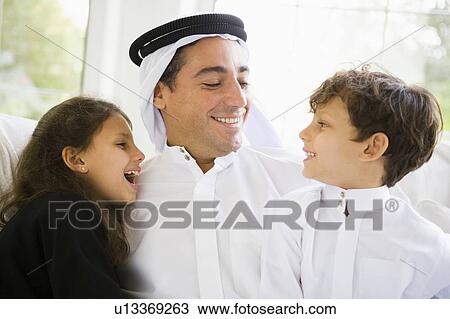 Stock Image - Father and two young children sitting in living room smiling (high key/selective focus). Fotosearch