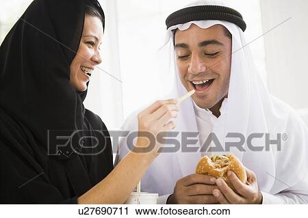 Stock Image - Couple eating fast food in living room and smiling (high key/selective focus). Fotosearch