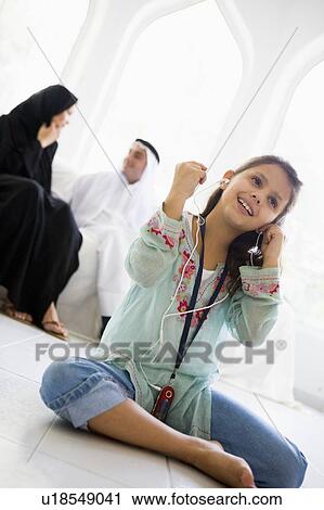 Stock Image - Daughter in living room listening to MP3 player and smiling with parents in background (high key/selective focus). Fotosearch