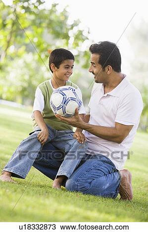 Stock Image - Father and son outdoors in park with ball smiling (selective focus). Fotosearch