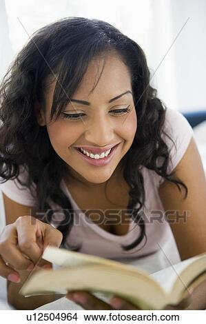 Picture - Woman in living room with book smiling (high key/selective focus). Fotosearch